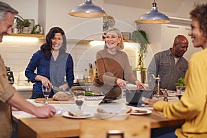 Group Of Mature Friends Meeting At Home Serving Food At Dinner Party And Drinking Wine Together