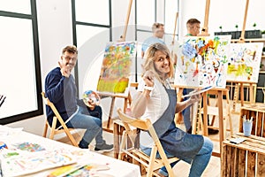 Group of middle age artist at art studio with a big smile on face, pointing with hand finger to the side looking at the camera