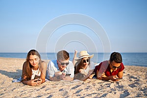 Group of millenials using smartphones laying together on beach towel near sea on summer sunset. Young people addicted by