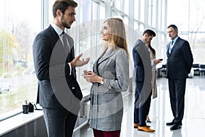 Group of modern business people chatting during coffee break standing in sunlit glass hall of office building.