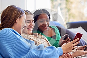 Group Of Multi-Cultural Teenage Girl Friends Snuggled Under Blanket Looking At Mobile Phones At Home