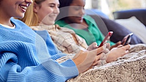 Group Of Multi-Cultural Teenage Girl Friends Snuggled Under Blanket Looking At Mobile Phones At Home