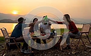 Group of multi-ethnic friends men and women sit on chair with party and camping together also cheers with bottle near lake in