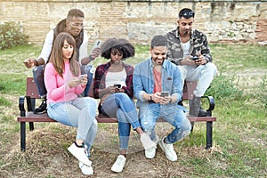 Group of multi-ethnic friends using their mobile phones while sitting on a bench together.