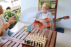 A group of multiethnic senior people playing guitar and sing a song after playing chess board with happy smiling face on sunny day