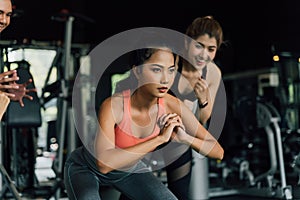 Group of people cheering on their Asian female friend doing squats with a weight plate in fitness gym. Working out