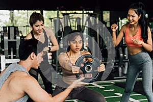 Group of people cheering on their Asian female friend doing squats with a weight plate in fitness gym. Working out