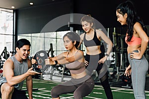 Group of people cheering on their Asian female friend doing squats with a weight plate in fitness gym. Working out