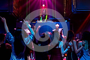 Group of people dance in disco night club to the beat of music from DJ on stage