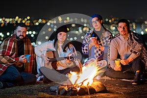 Group of people having fun sitting near bonfire outdoors at night playing guitar, singing songs and talking happily together