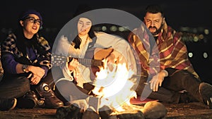 Group of people having fun sitting near bonfire outdoors at night playing guitar, singing songs and talking happily together.