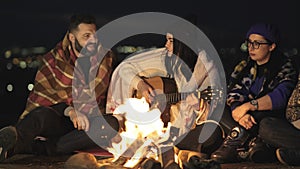 Group of people having fun sitting near bonfire outdoors at night playing guitar, singing songs and talking happily together.