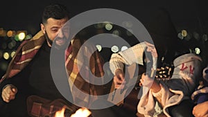 Group of people having fun sitting near bonfire outdoors at night playing guitar, singing songs and talking happily together.