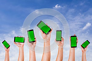 group of people& x27;s hands holding phones with green screen and rising them up against a blue sky