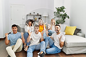 Group of people sitting on the sofa and floor at home celebrating victory with happy smile and winner expression with raised hands