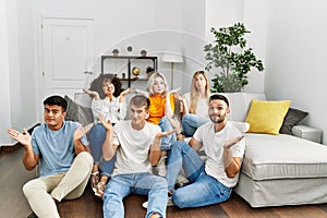Group of people sitting on the sofa and floor at home clueless and confused expression with arms and hands raised