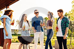 Group of people stand around the grill, chatting, drinking and eating. Young friends are having a barbecue party