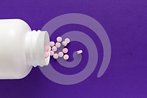 Group pink tablets. Capsules spilling out of white bottle. Purple background.