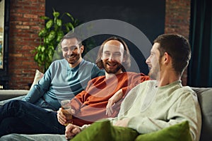group portrait of cheerful friends watching football game, soccer match in living room on sofa and drinking alcohol