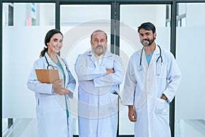 Group portrait of diverse male and female doctors in white medical uniforms standing in hospital corridor smiling and looking at
