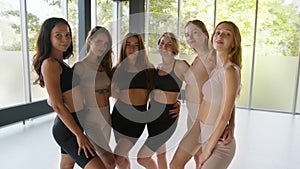 Group portrait of flexible ladies posing together at modern light room, embracing and smiling to camera, tracking shot