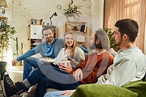 Group portrait. Guys and girl sitting on couch in living room and discussing while watching comedy series, TV show on