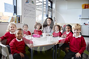 Group portrait of infant school teacher and kids sitting at table in a classroom looking to camera smiling, front view