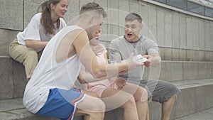 Group of positive Caucasian young men and women sitting on urban stairs as cheerful guy coming up to them with break