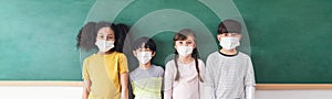 Group of positive elementary school kids wearing medical face masks to prevent disease standing in front of chalkboard in