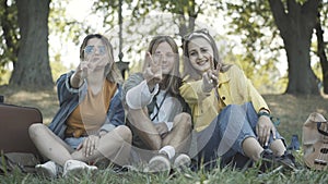 Group of positive hippies showing peace gesture and smiling. Happy relaxed Caucasian friends sitting on green summer