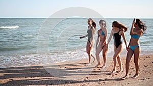 Group of pretty female friends walking by the sea on sandy tropical beach in swimsuits. Interracial women in bikini