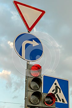 A group of road signs and a traffic light with an additional section at the intersection against the sky. A red traffic light and