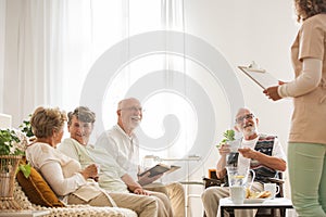 Group of senior nursing home pensioners sitting together at common living room listening to young nurse