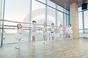 Group of seven little ballerinas standing in row and practicing ballet using stick on the wall