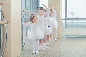 Group of seven little ballerinas standing in row and practicing ballet using stick on the wall