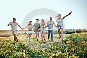 Group of six friends running down meadow, jumping, cheering and smiling. Friends having fun