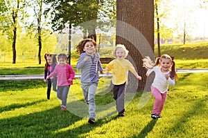 A group of small happy children run through the park in the background of grass and trees.