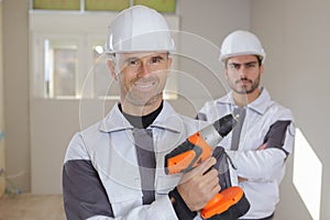 Group smiling builders in hardhats with electric drill indoors