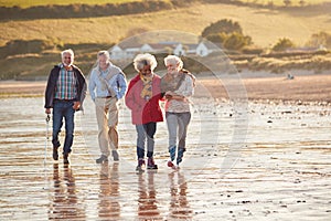 Group Of Smiling Senior Friends Walking Arm In Arm Along Shoreline Of Winter Beach