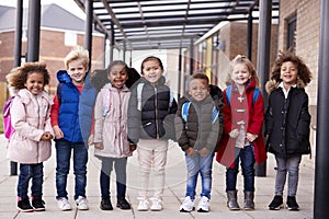 A group of smiling young multi-ethnic school kids wearing coats and carrying schoolbags standing in a row in walkway outside their