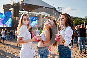 Group of smiling young women with sunglasses enjoy summer music festival on beach holding colorful drinks, casual