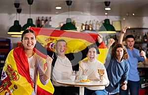 Group of sports supporters cheering for favorite team with flag of Spain while watching match game on TV in bar