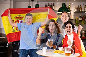 Group of sports supporters cheering for favorite team with flag of Spain while watching match game on TV in bar