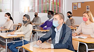 Group of students in protective mask listening attentively to teacher explaining material in the classroom