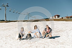 Group of stylish tourists and a dog sitting in the snow in spring in a mountain resort and posing for the camera