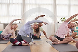 Group With Teacher Sitting On Exercise Mats Stretching In Yoga Class Inside Community Center