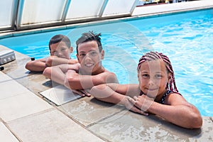 Group of teenage children enjoy swimming in pool, two boys and one girl looking at camera