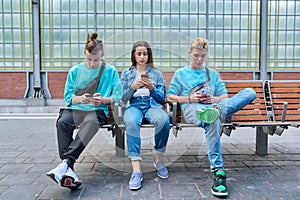 Group of teenage friends using smartphones sitting on chairs on train station platform