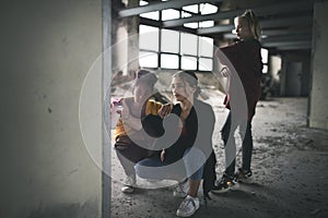 Group of teenagers girl gang indoors in abandoned building, using spray paint on wall.