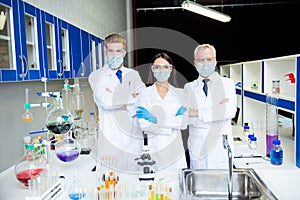 Group of three assistants doing vaccine experiment in lab all wearing labcoats gloves glasses gauze masks folded arms